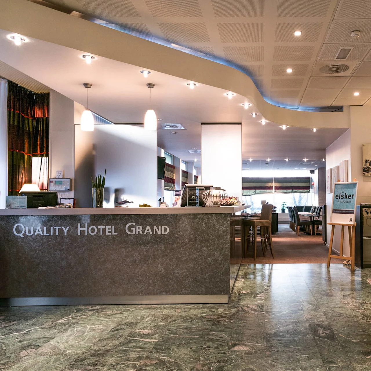 Reception desk with "QUALITY HOTEL GRAND" inscription; lounge area with armchairs and table. Illuminated interior; marble floor; poster with "elsker" text on easel, lamps and wall art visible.