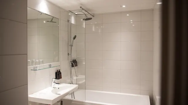Ett badrum innehåller ett badkar med en dusch ovanpå, ett vitt handfat med flaskor och en stor spegel, omgivet av vita kaklade väggar och mjuk belysning.