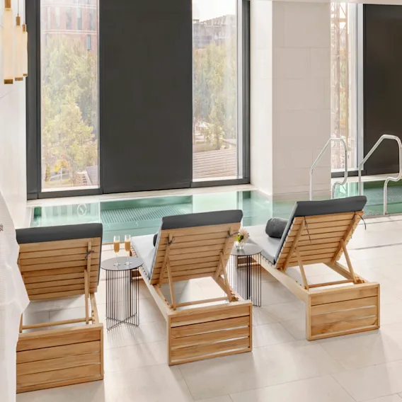 Sunchairs with cushions face an indoor swimming pool, adjacent to large windows. A small table holds glasses of champagne. A robe hangs nearby with text "elements spa."