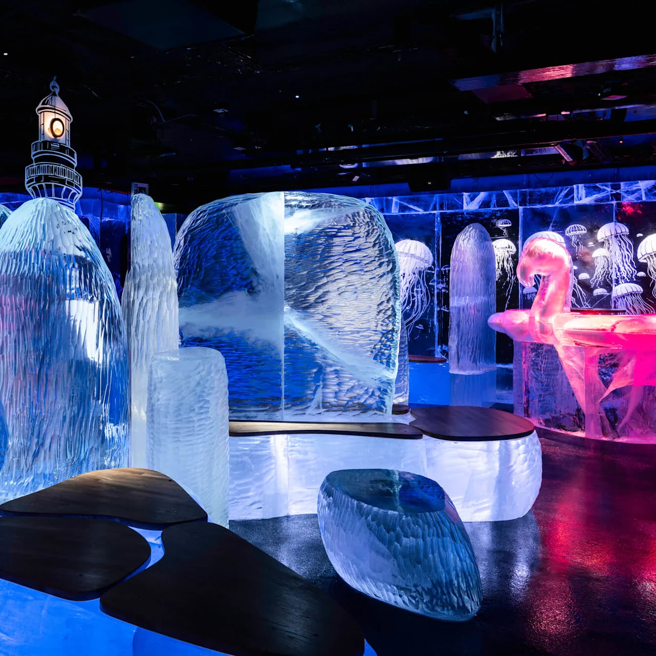 Ice sculptures are illuminated in blue and pink, featuring a flamingo and jellyfish designs. Wooden benches are placed amidst the icy structures in a dimly lit environment.