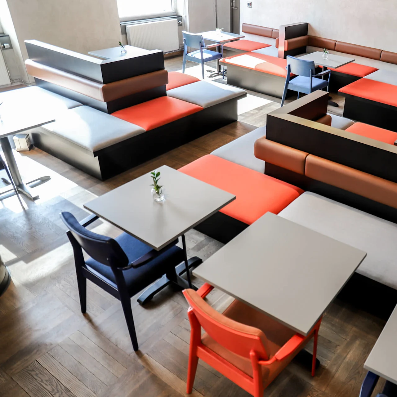 Tables and chairs arranged in a grid, surrounded by red and gray cushioned benches, create a modern seating area in a well-lit room with wooden flooring.