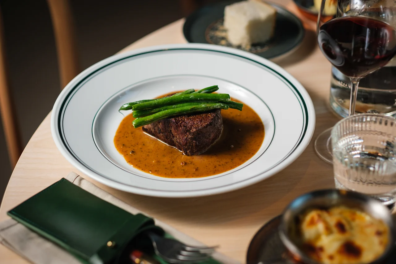 En biff toppas med gröna bönor och serveras i en brun sås på en vit tallrik, omgiven av ett glas rödvin, bröd och bestick på ett träbord.