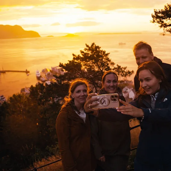 En gruppe venner tar en selfie med en vakker solnedgang over en kystby og vann i bakgrunnen.