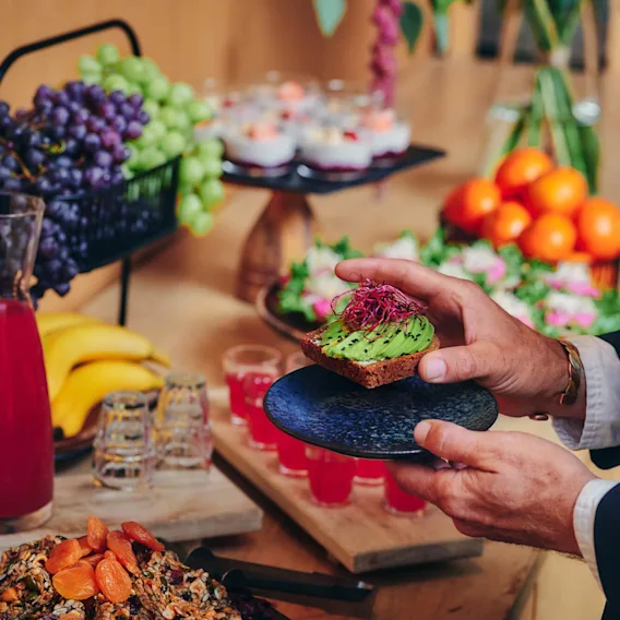 En person i kostym håller en tallrik med avokadotoast och står vid ett bord fullt med frukt, nötter och drycker.