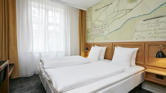 Et lyst hotelværelse med to enkeltsenge, hvidt sengetøj og et stort vindue med tynde gardiner. Et gammelt kortmaleri pryder væggen.
