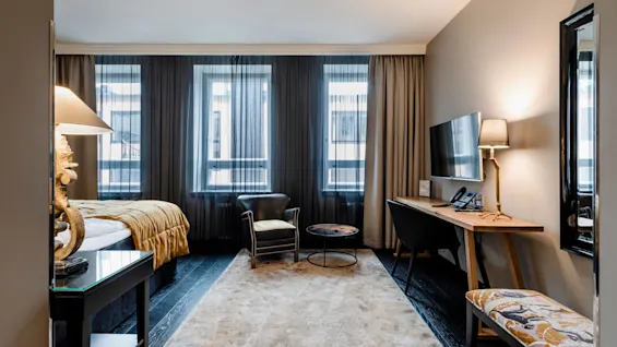 Et moderne hotellrom med seng, lenestol og skrivebord med lampe og TV. Store vinduer med lette gardiner skaper en lys, koselig atmosfære.
