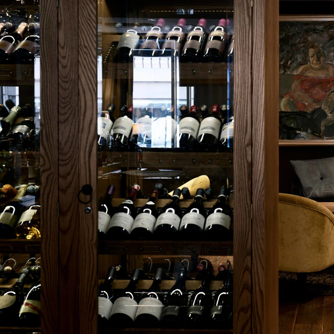 Wine bottles are stored in a wooden cabinet, with a cozy seating area beside it. Two yellow armchairs face a table with a chess set, lit by a floor lamp.