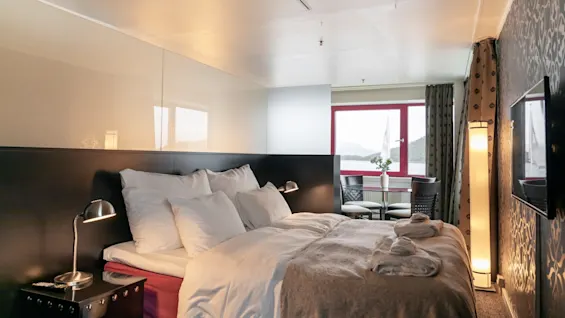 Viihtyisä hotellihuone, jossa on mukava sänky, ruokailutila ikkunan äärellä ja luonnonkaunis näkymä järvelle ja vuorille.