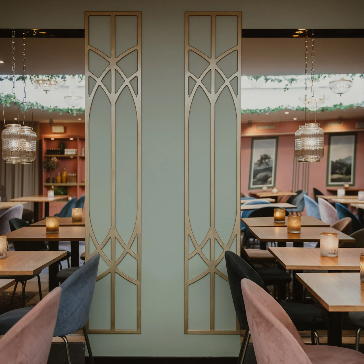 Borde med lyserøde og blå stole er dækket med lys i en stilfuld, blødt oplyst restaurant. Dekorative skillevægge med geometriske mønstre adskiller siddeområderne og skaber hyggelig stemning.