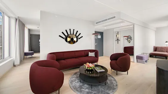 A burgundy seating arrangement with two chairs and a sofa surrounds a round coffee table and rug in a modern, spacious living room with large windows and a wall mirror decoration.