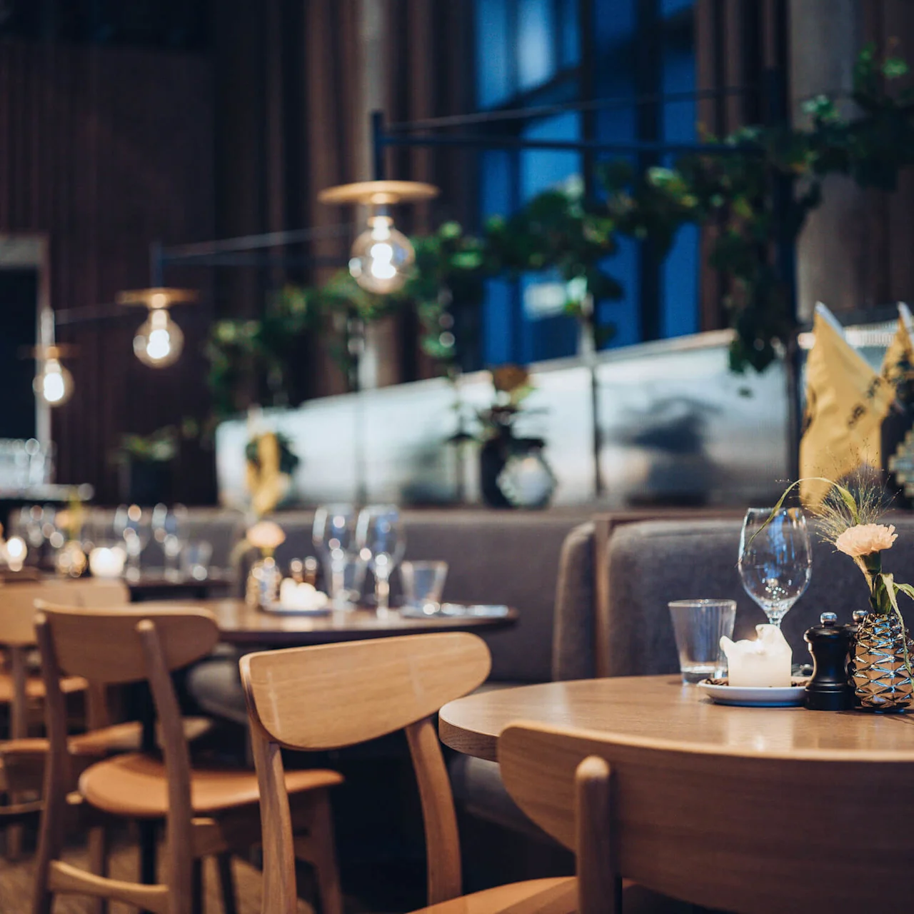 Wooden dining tables are set with glasses and candles, surrounded by chairs in a softly lit, elegant restaurant with hanging lights and decorative plants on a cozy evening.
