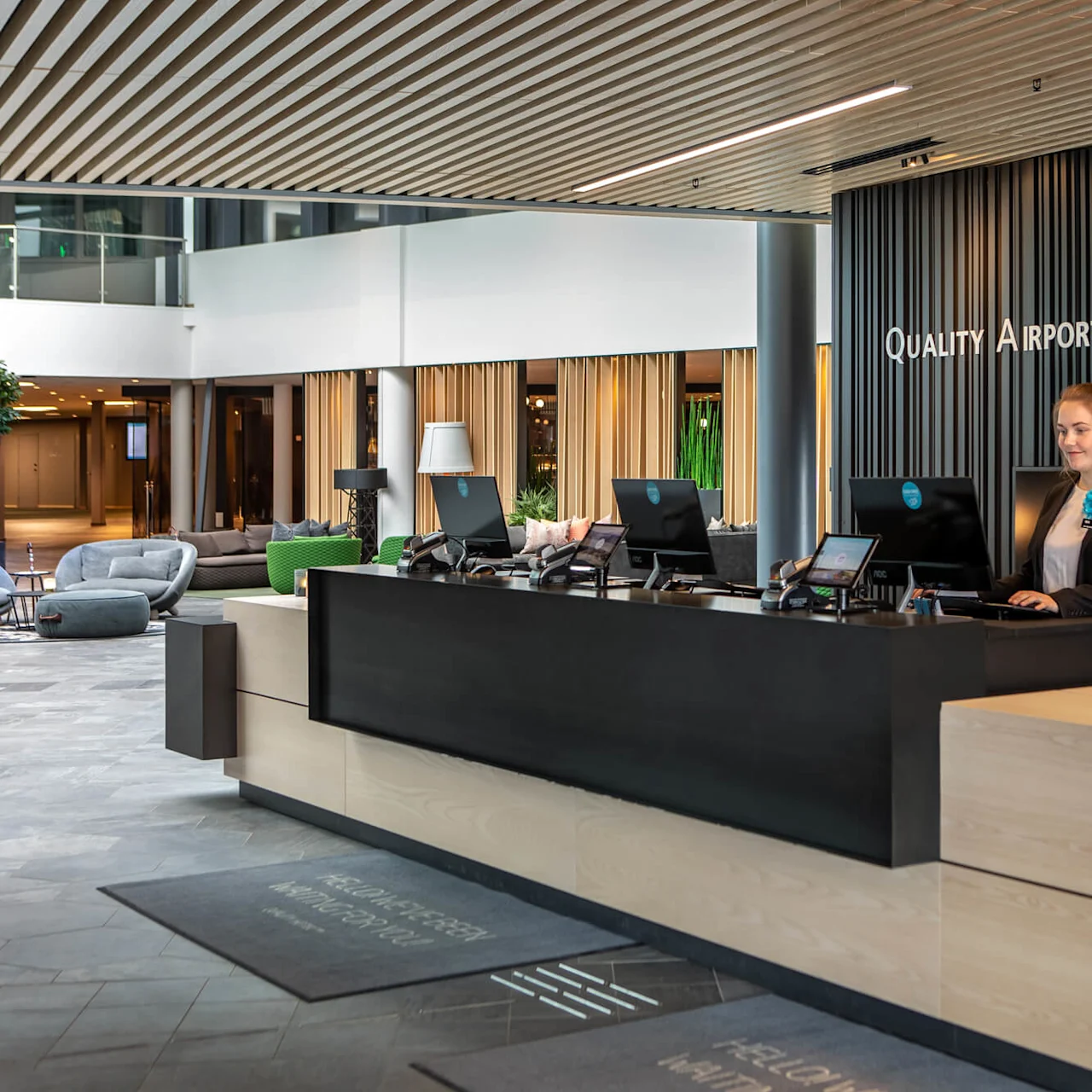 A reception desk with a receptionist working on a computer, situated in a modern hotel lobby. Text: "QUALITY AIRPORT HOTEL GARDERMOEN."