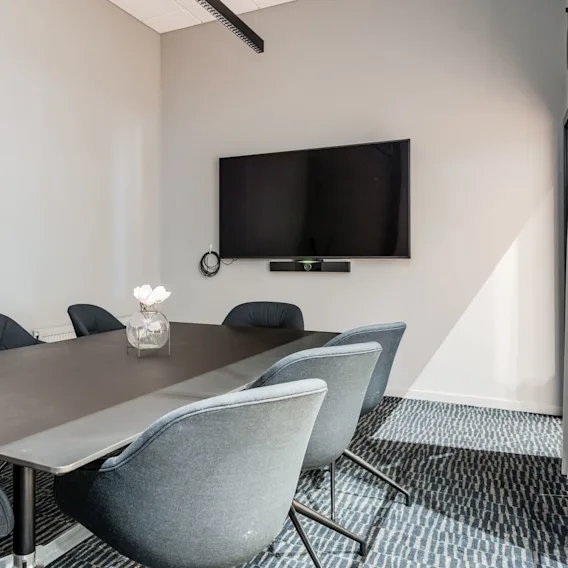A modern conference room with a black table and six gray chairs. A large television is mounted on the wall, with a vase of white flowers on the table.