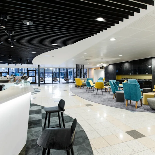 Valoisa, moderni lounge, jossa värikkäät keltaiset ja siniset tuolit matalien pöytien ympärillä matolla, vierekkäin tyylikkään valkoisen baaritiskin kanssa, tumman katon alla.