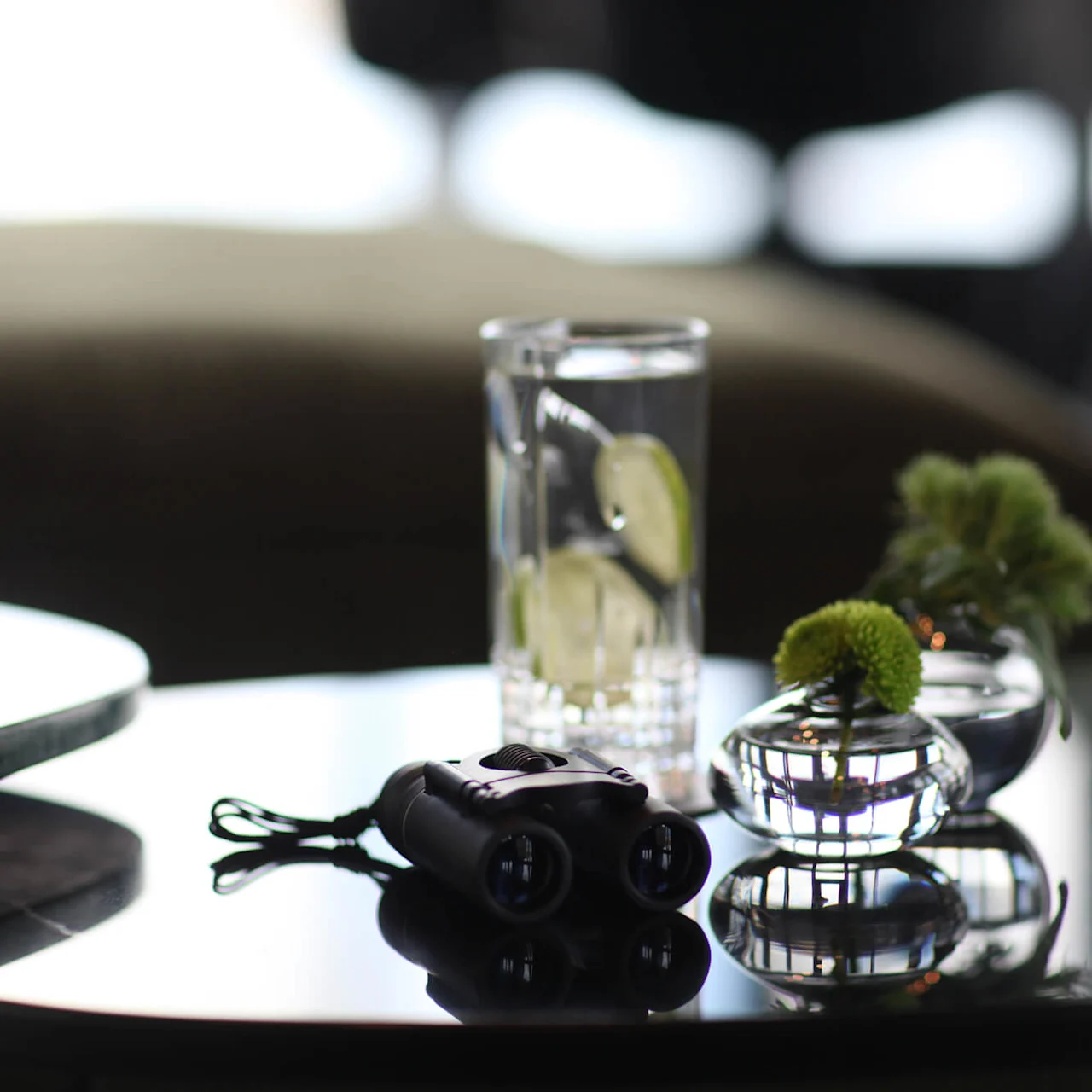 Binoculars rest on a reflective table beside a glass of water with lime slices and two small vases with green flowers, against a blurred indoor background.