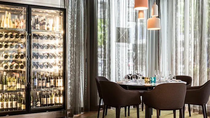 A wine fridge displaying bottles stands beside a round table with set wine glasses, plates, and cutlery. Soft pendant lights illuminate the modern, elegant dining room with sheer curtains.
