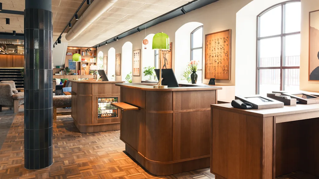 Interiør av en hotellobby med resepsjonsdisker i tre, mønstret gulv og naturlig lys fra buede vinduer.