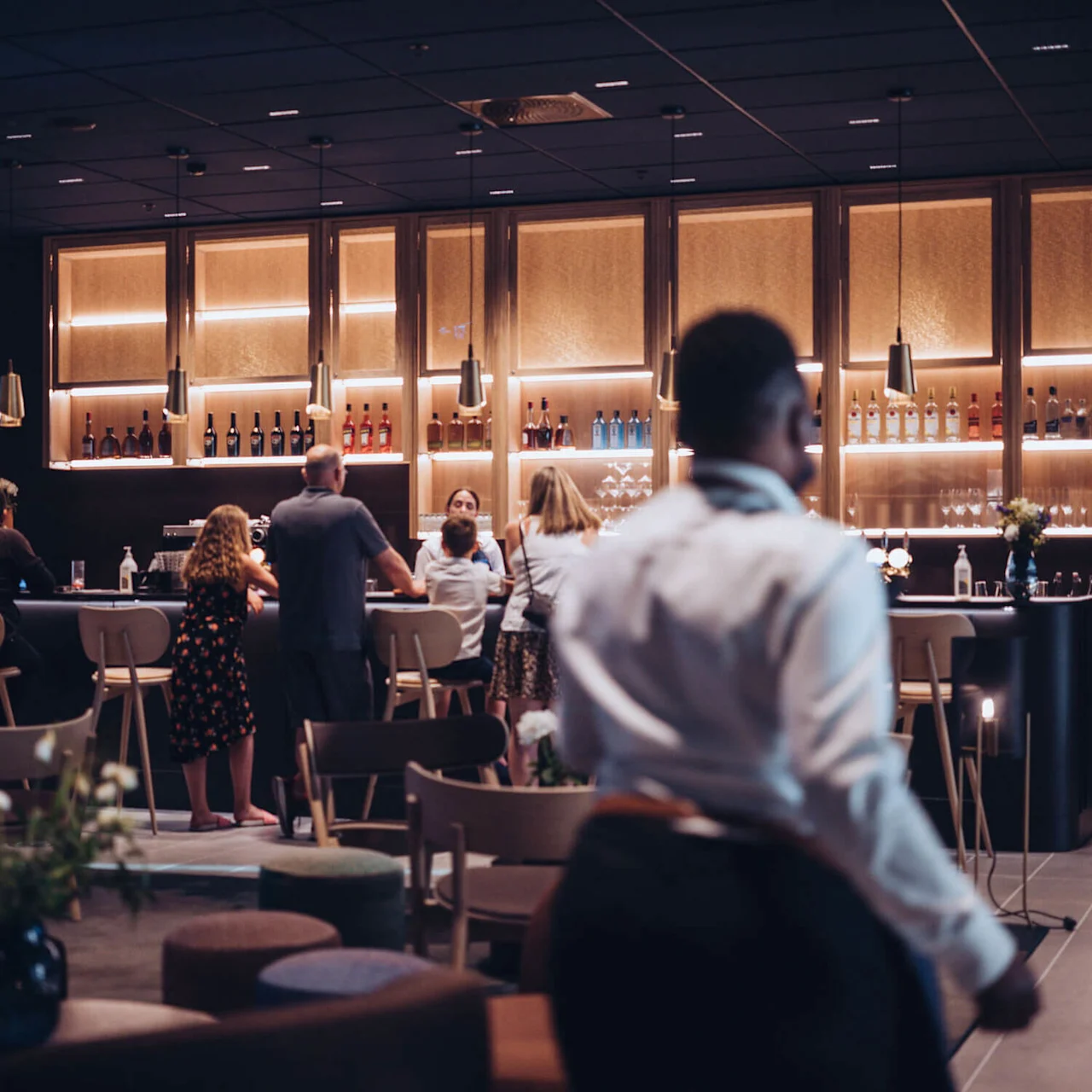 English: People interact at a modern bar, with bottles on backlit shelves. A person in a white shirt walks in the foreground. Soft lighting creates a welcoming atmosphere.