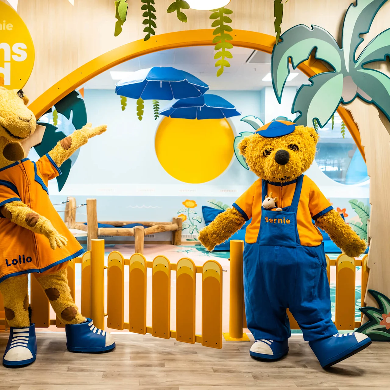 Giraff- og bjørnemaskotter, klædt i orange og blå outfits, står muntert i et børnelegeområde med farverige tropiske dekorationer. Tekst: "Lollo & Bernie, Solens land".