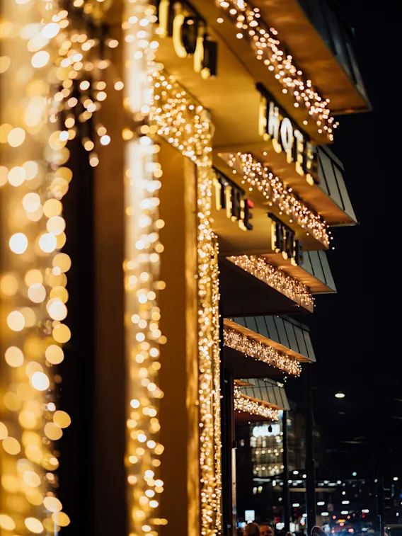 Julelys dekorerer bygningens markiser om natten; guldskrift stave "HOE" og "THE." Fodgængere går nedenunder, midt i en travl by gade. Bygninger oplyses i baggrunden.