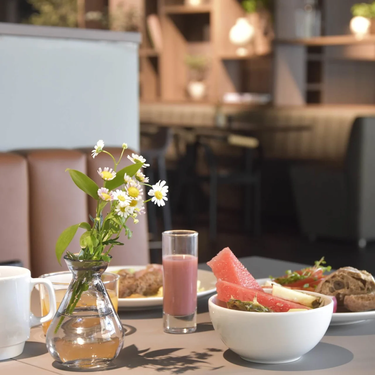 A vase with white daisies sits on a table with breakfast items; a smoothie, watermelon, and bread. The background features a cozy, plant-filled cafe interior.
