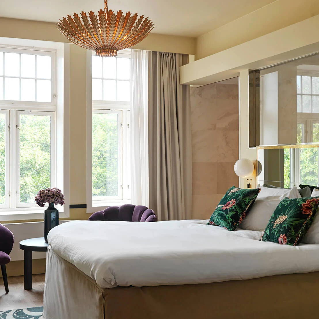 A neatly made bed with floral pillows rests in a stylish bedroom. Two purple chairs, a small table with flowers, and large windows allow natural light to fill the room.