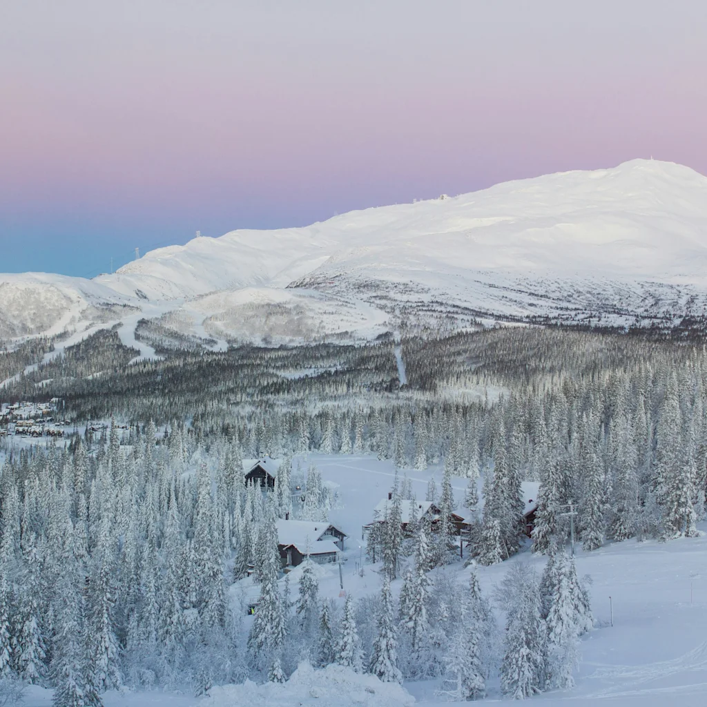 Ett fridfullt vinterlandskap med snötäckta berg, en tät tallskog och små stugor under en rosa och blå himmel.