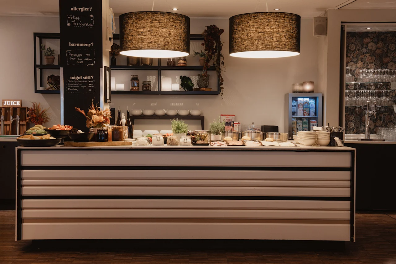 En veloplyst buffetdisk med forskellige madmuligheder, drikkevarer og skilte, der lyder allergier, barnmeny, nagot sott og JUICE.
