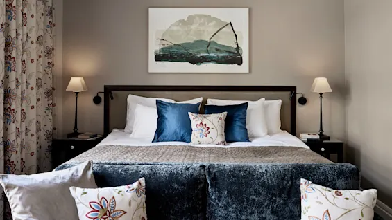 En sirligt redt seng med blå og blomstrede puder står mod en beige væg. Ovenfor, abstrakt kunst; to sideborde med lamper flankerer sengen, omgivet af mønstrede gardiner.