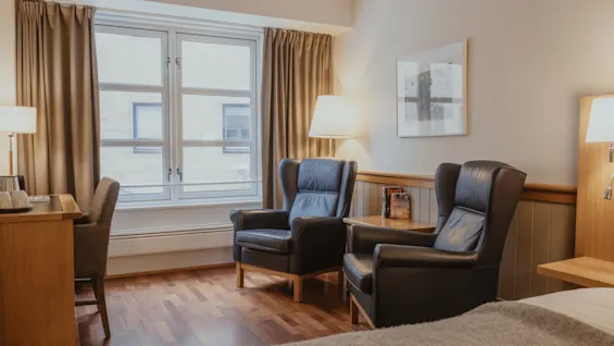 A cozy hotel room with a comfortable bed, two armchairs, a desk, and a large window offering natural light.