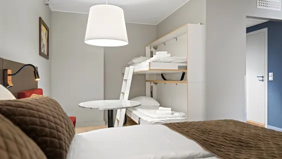 En dobbeltseng med brune tæpper står over for en køjeseng mod en lysegrå væg. Et rundt bord og en lampe tager pladsen i midten; håndklæder ligger på sengene.