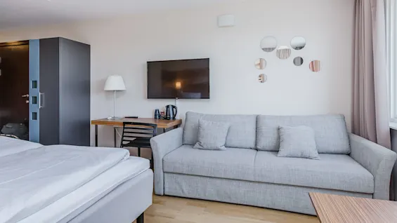 Kodikas hotellihuone, jossa on siisti sänky, harmaa sohva ja seinälle asennettu televisio. Lähellä on pöytä, jossa on lamppu ja vedenkeitin. Pyöreät peilit koristavat valkoista seinää.