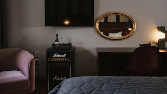 TV monterad på en vägg, ovanför en Marshall-förstärkare med en vinflaska och glas; bredvid speglas sängkläder i en oval spegel, och en mjukt belyst lampa står på ett skrivbord.