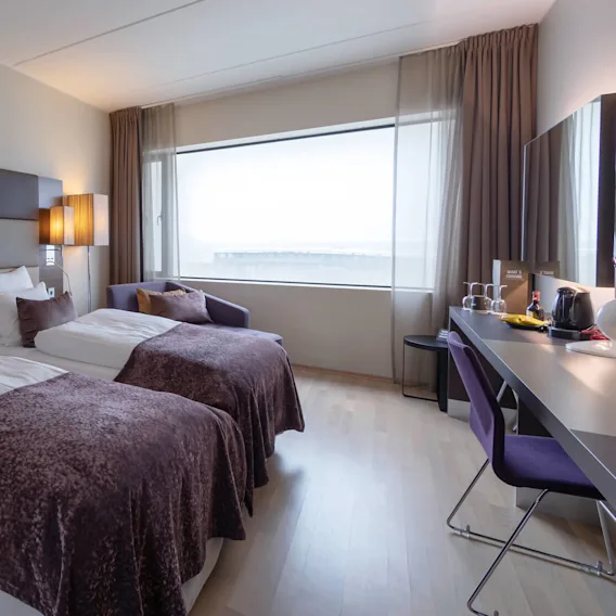 To prydelig oppredde enkeltsenger med brune tepper står side om side i et moderne hotellrom med et stort vindu, stoler og et skrivebord med vannkoker og kopper.