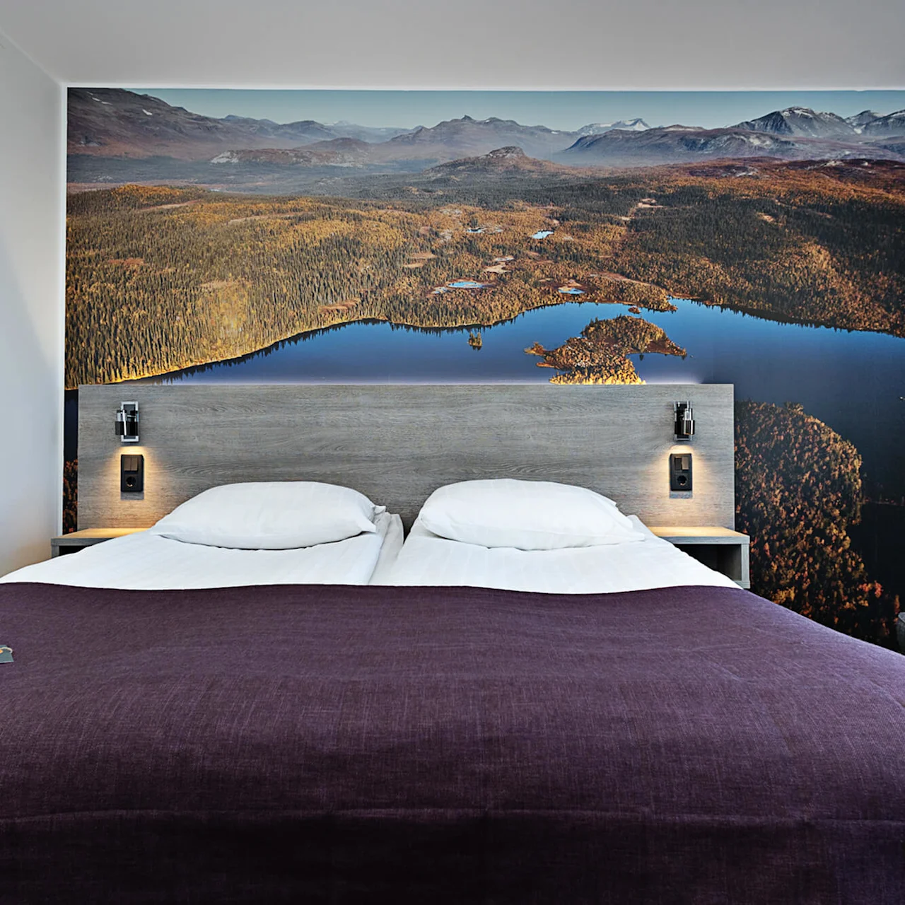 En prydligt bäddad dubbelsäng står mot en sänggavel med en naturtrogen landskapstapet. Mjuk belysning och ett mörklila överkast fullbordar den mysiga hotellrumsatmosfären.