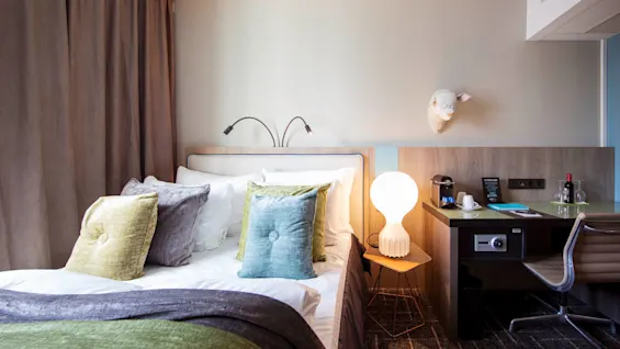Bed with colorful pillows next to a small table with a glowing lamp in a modern hotel room. A sturdy desk holds a safe, chair, coffee cup, and wine bottle.