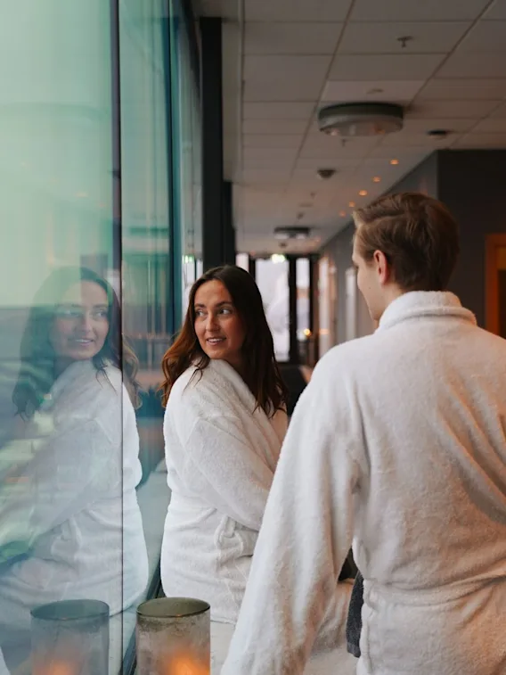 A man and woman wearing white bathrobes standing near a large window in a cozy spa relax area.