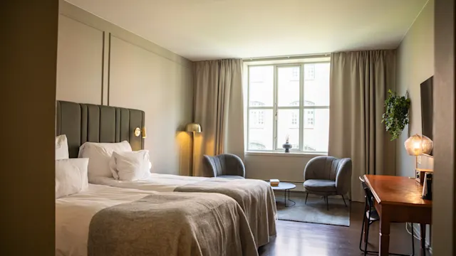 En seng med gråe tepper og hvite puter. To stoler og et bord ved et stort vindu. Myk belysning og nøytrale toner skaper en hyggelig hotellrom-atmosfære.