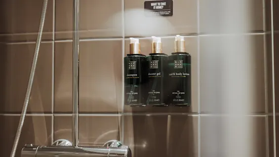 Tre gröna flaskor märkta "shampoo," "shower gel," och "milk & body lotion" på en duschvägg. Text ovan lyder: "WANT TO TAKE IT HOME? PLEASE BUY IT AT THE FRONT DESK."