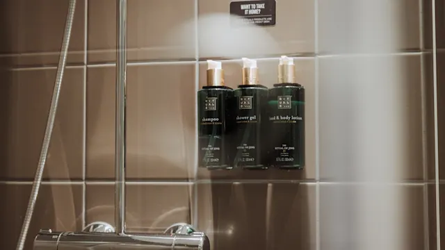 Tre gröna flaskor märkta "shampoo," "shower gel," och "milk & body lotion" på en duschvägg. Text ovan lyder: "WANT TO TAKE IT HOME? PLEASE BUY IT AT THE FRONT DESK."