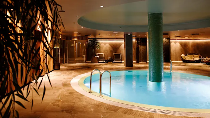 A round indoor pool with a central pillar, surrounded by lounge chairs and plants, in a warmly lit spa setting.