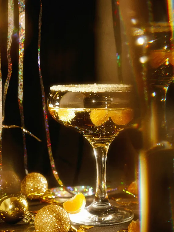 Ett champagneglas med sockrad kant och mandarinklyftor omgivet av glittrande juldekorationer i guld.