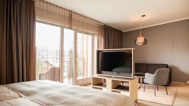 Sengen vender mot en stor TV på et stativ, med en grå lenestol og sofa i nærheten. Stort vindu med gjennomsiktige gardiner slipper inn naturlig lys og skaper en koselig atmosfære.