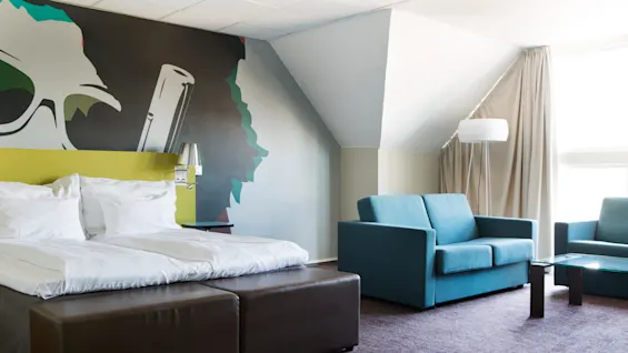 A double bed with white linens stands beside a colorful mural depicting large glasses. A blue couch and glass table are in a softly lit room with beige curtains.