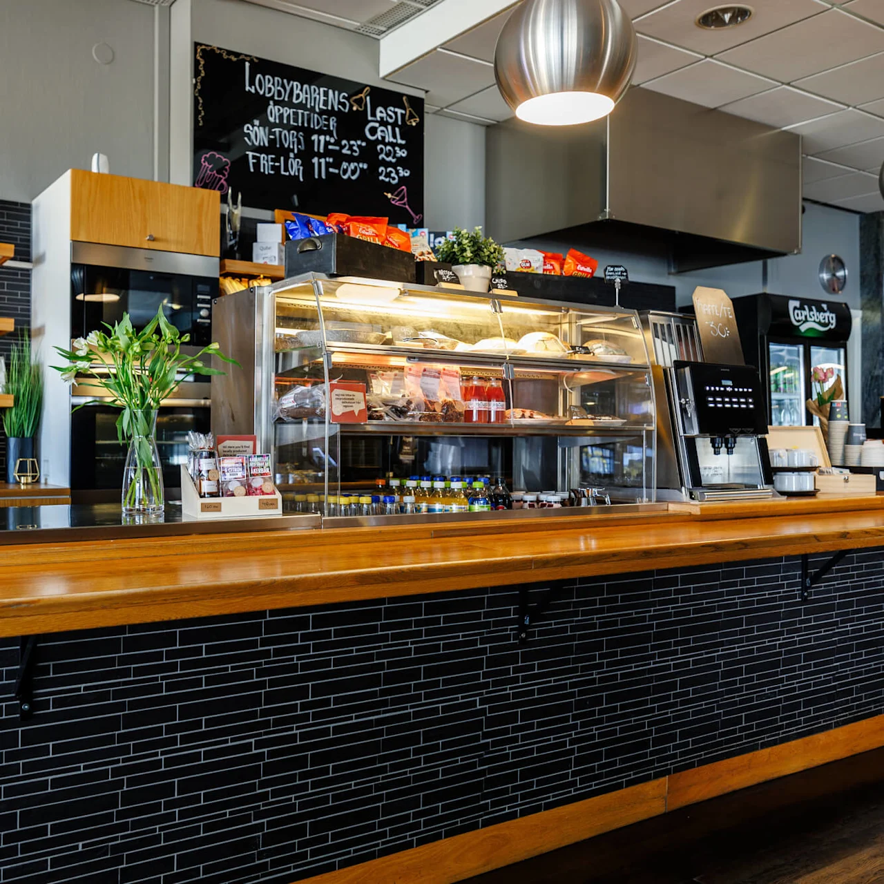 Ett träkafédiska visar snacks under ett glashölje; en meny tavla visar "LOBBYBARENS ÖPPETTIDER, SÖN-TORS 11-23, FRE-LÖR 11-00, LAST CALL 22:30". Hyllor håller glas; hängande lampor belyser.