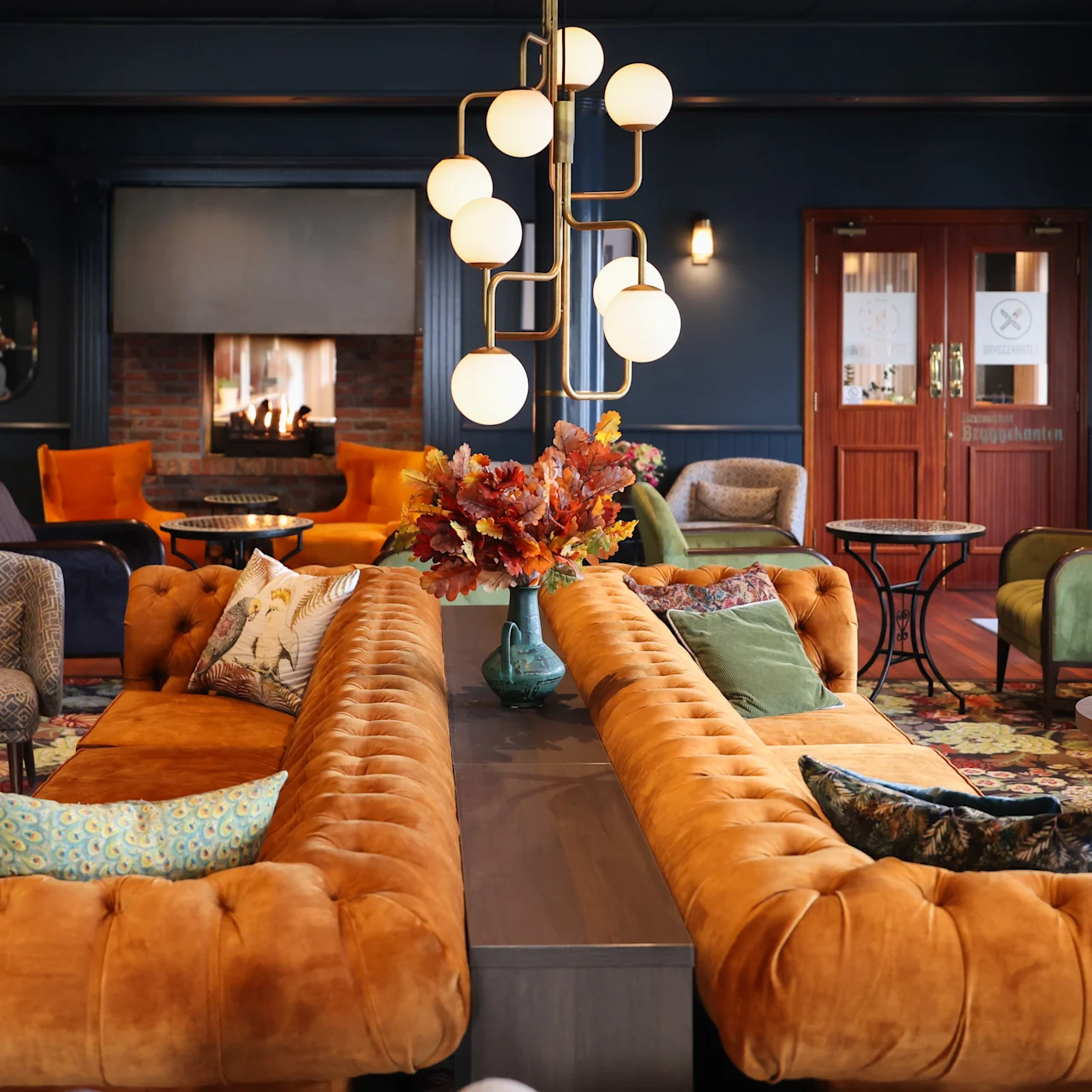 A plush, orange sectional sofa with cushions is centered in an elegant, warmly lit room. Beyond, a cozy fireplace is surrounded by assorted seating, including chairs and tables arranged on a patterned rug.