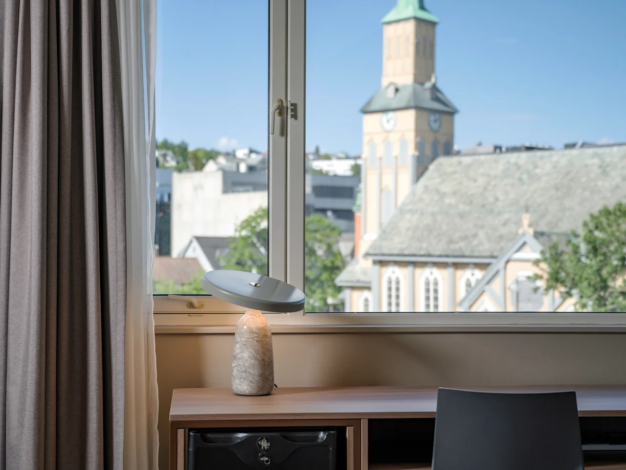 En skrivebordslampe hviler på et trebord foran et vindue med gardiner, med udsigt over en bysilhuet, der inkluderer et kirketårn under en blå himmel.