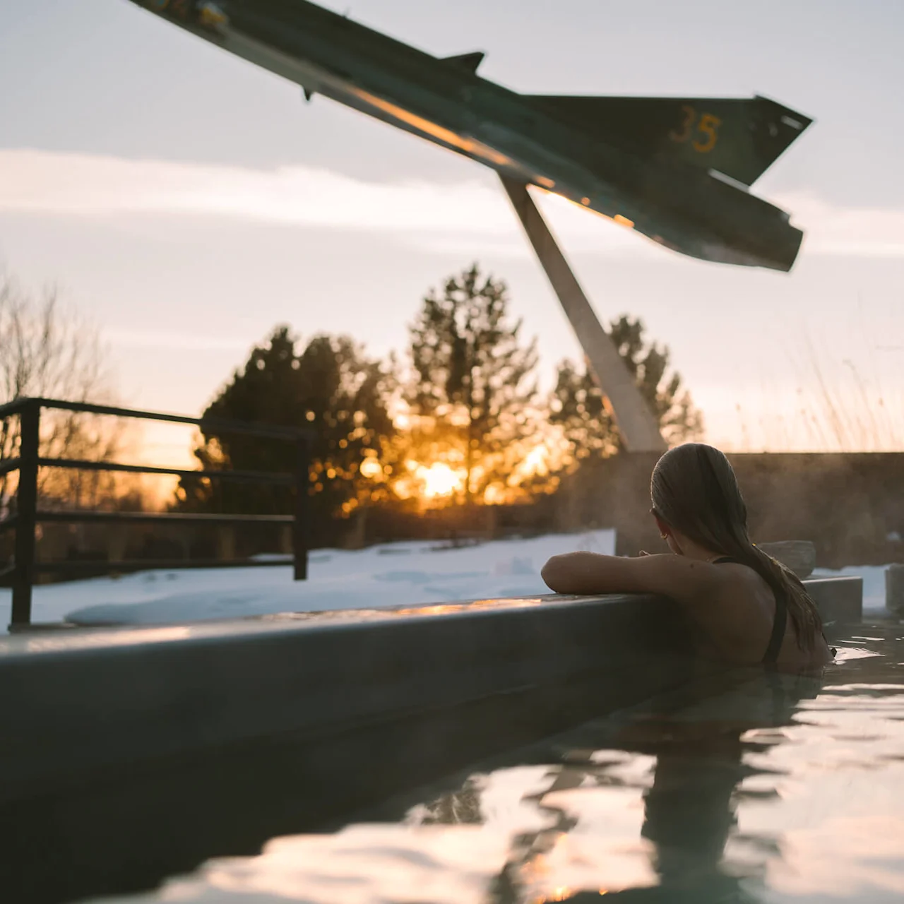 En person kopplar av i en utomhusjacuzzi vid solnedgången, omgiven av snö. En stor flygplansskulptur märkt "35" finns i närheten.