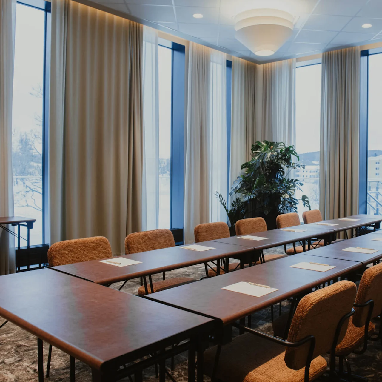 Bord arrangerade i en U-form visar papper och pennor, omgivna av stolar i ett modernt konferensrum med stora fönster, beige gardiner och en krukväxt i hörnet.