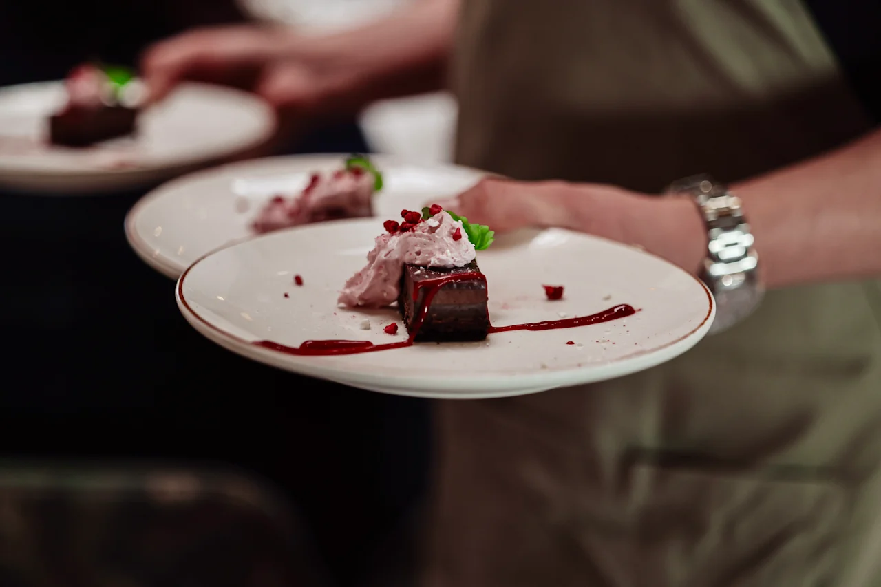 A server carries two white plates, each holding a chocolate dessert topped with berry sauce and garnished with cream and mint, in a dimly lit dining setting.